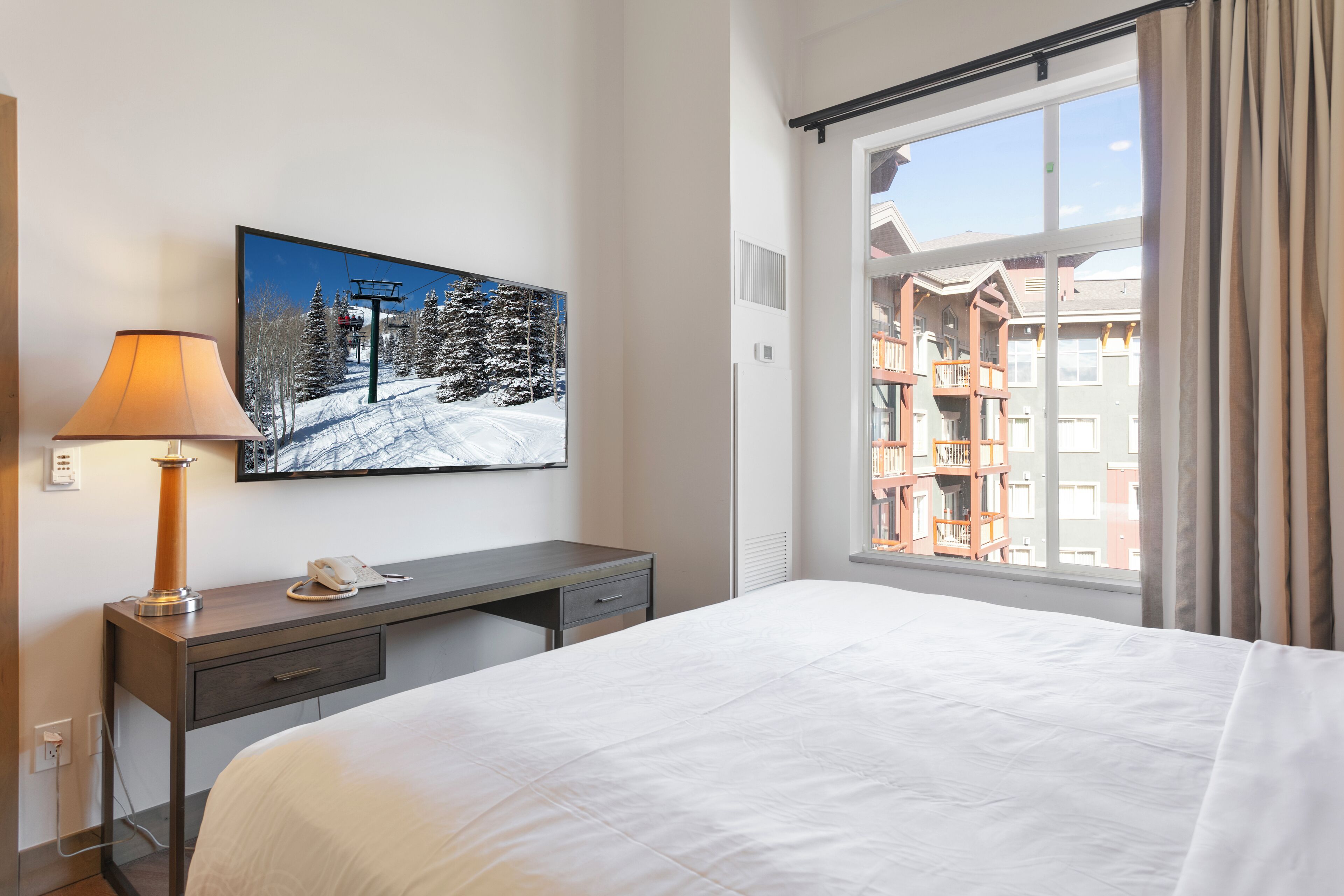 Hotellihuone, jossa on sänky, työpöytä lampun kanssa, puhelin ja suuri ikkuna, josta on näkymä rakennukseen.
