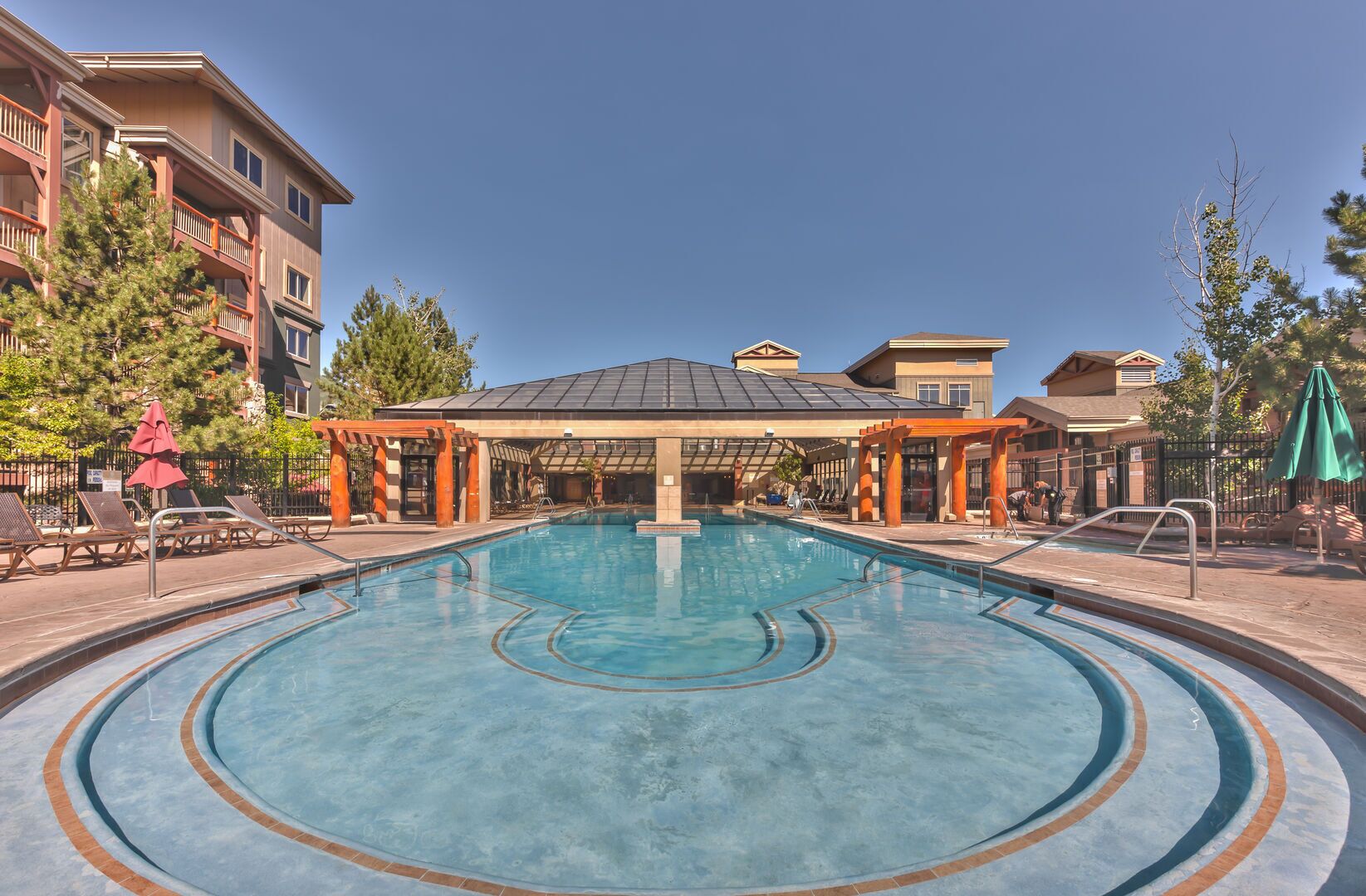 A swimming pool with a sunken center, surrounded by lounge chairs and umbrellas, with buildings in the background.