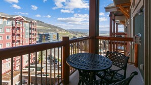 Un balcon avec une table ronde et des chaises, d’où l’on peut admirer un paysage urbain composé d’immeubles à plusieurs étages et, au loin, une chaîne de montagnes.