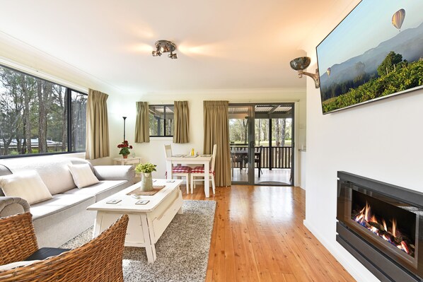 A spacious living room with a sofa, coffee table, and fireplace. There is a dining area with a table and chairs, and a view of the outdoors through large windows.