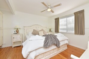A bedroom with a bed, two bedside tables, a ceiling fan, a window with curtains, and a wooden floor.