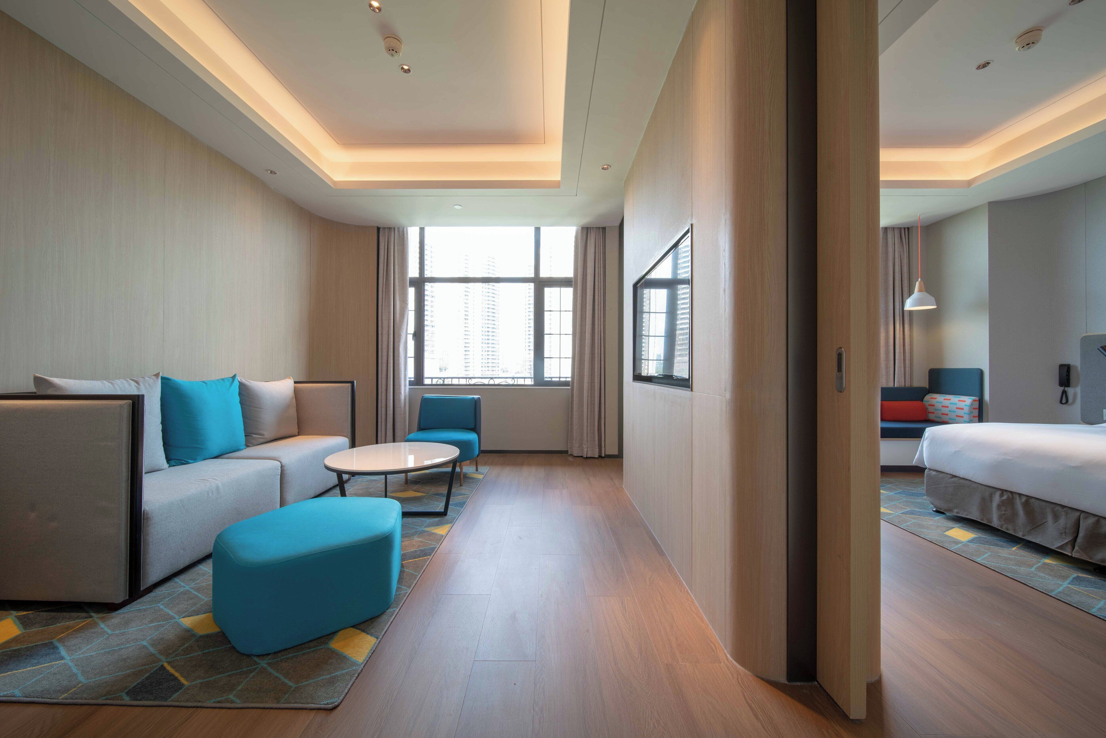 A modern hotel room with a sofa, a small round table, a bed, and a large window providing natural light.