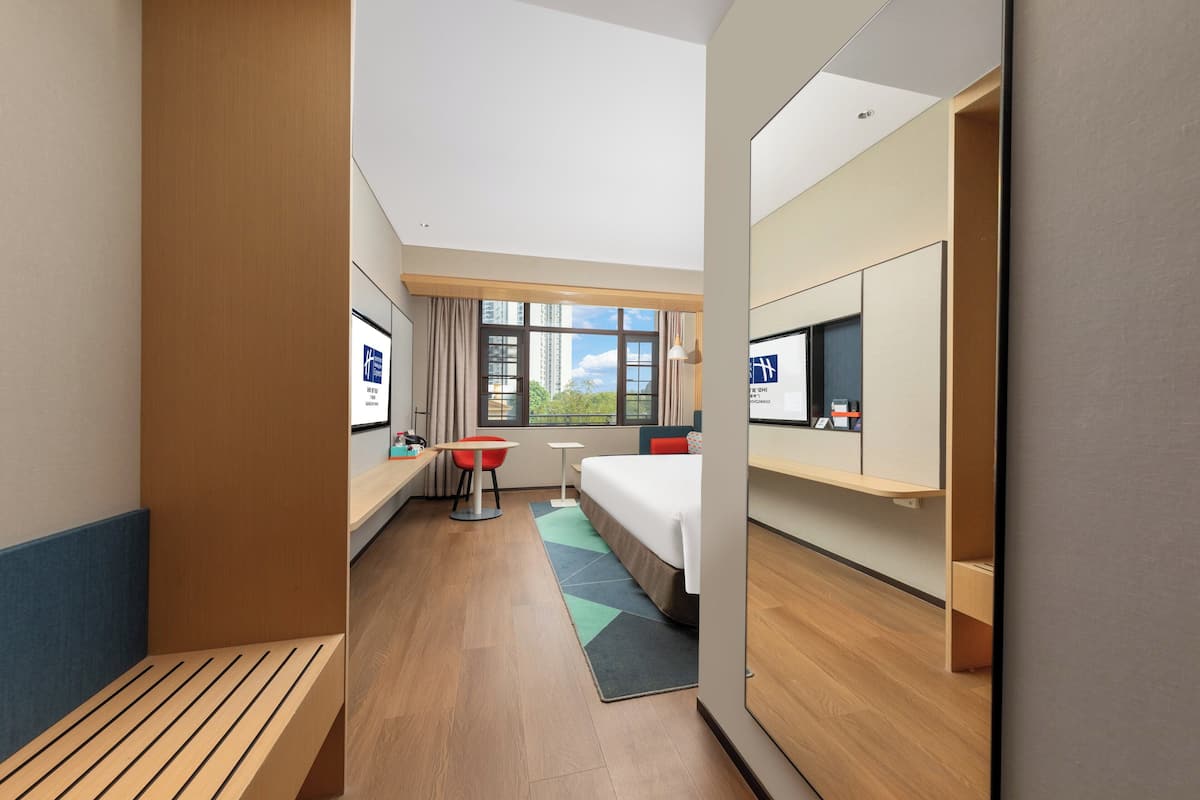 A hotel room with a bed, a desk, a chair, a TV, and a window with a view of buildings.