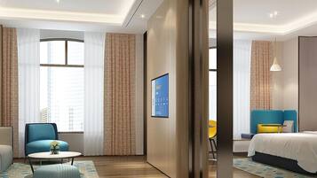 A modern hotel room with a large bed, a seating area with a chair and a small table, and a large window providing natural light.