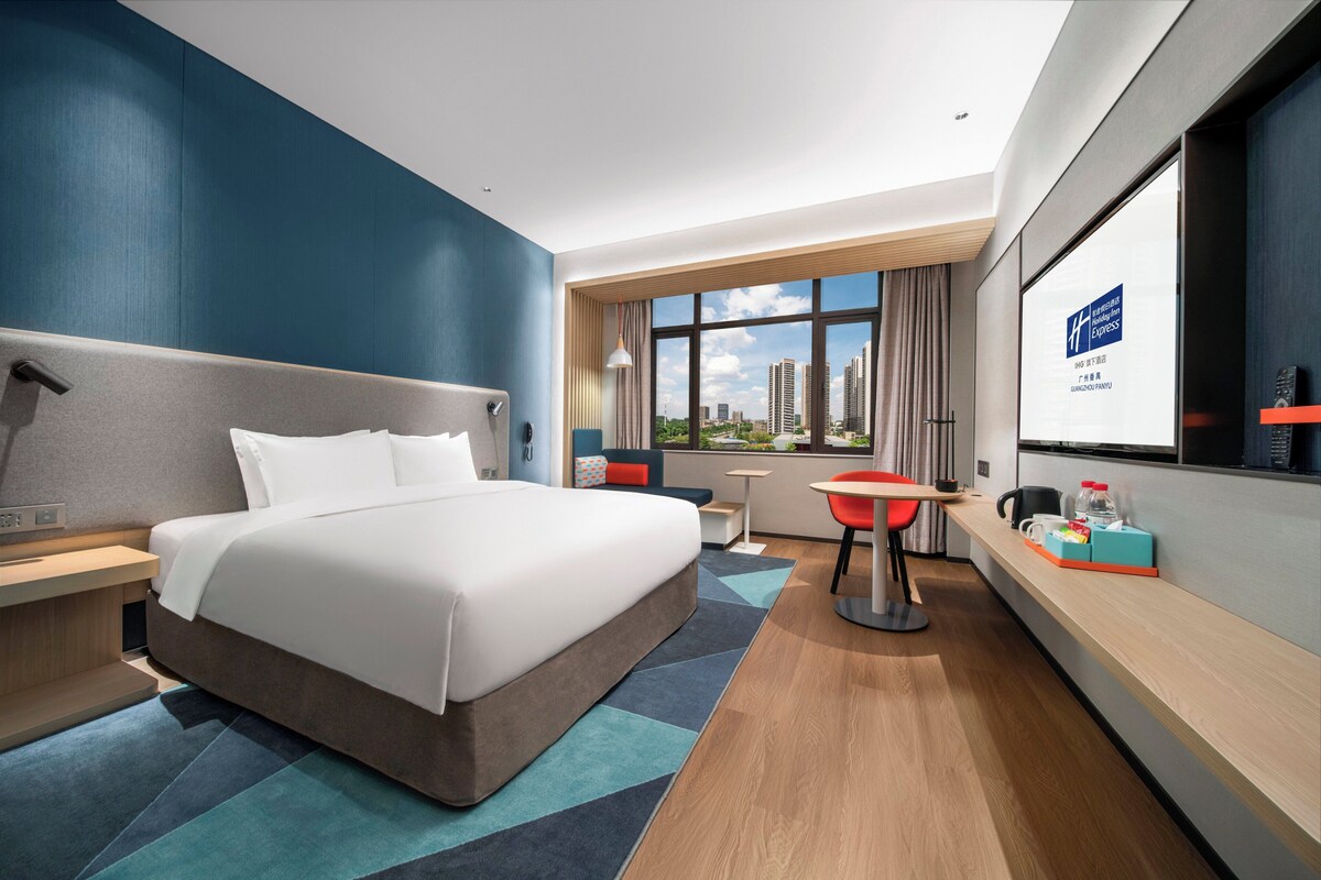 Een moderne hotelkamer met een groot bed, een bureau, een stoel en een uitzicht op de stad door grote ramen.