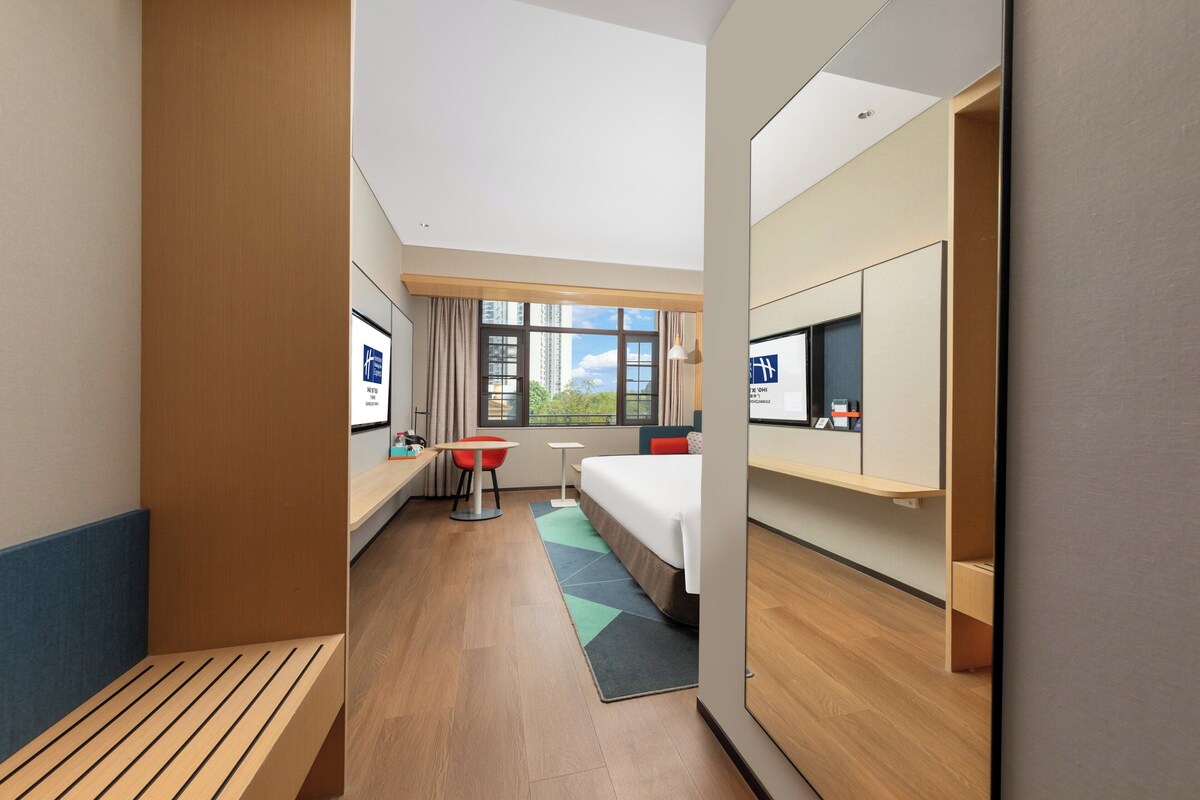 Een hotelkamer met een bed, een bureau, een stoel, een televisie en een raam met uitzicht op gebouwen.