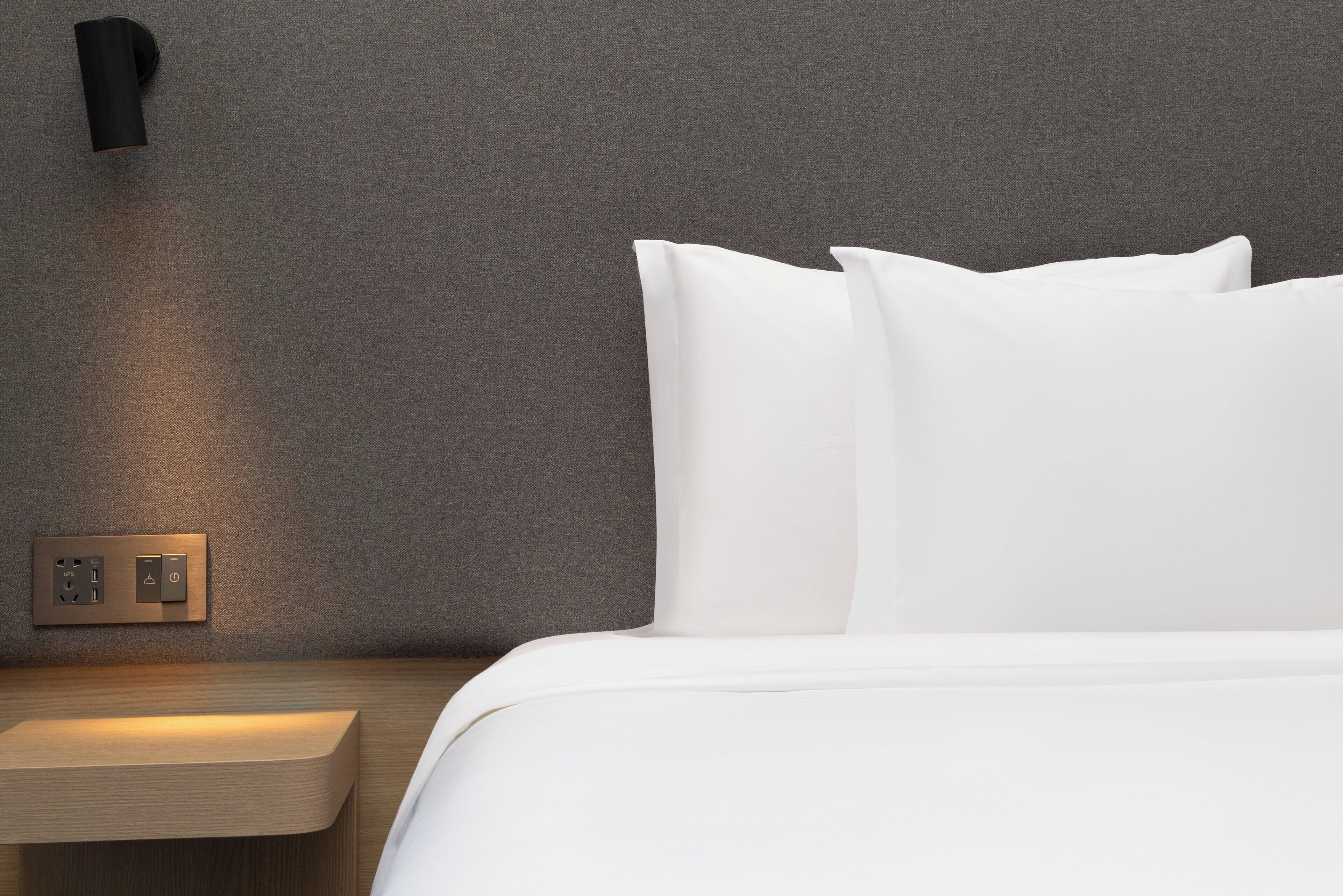 Una cama bien hecha con sábanas blancas, una mesita de noche con interruptor de luz y una lámpara fijada a la pared.