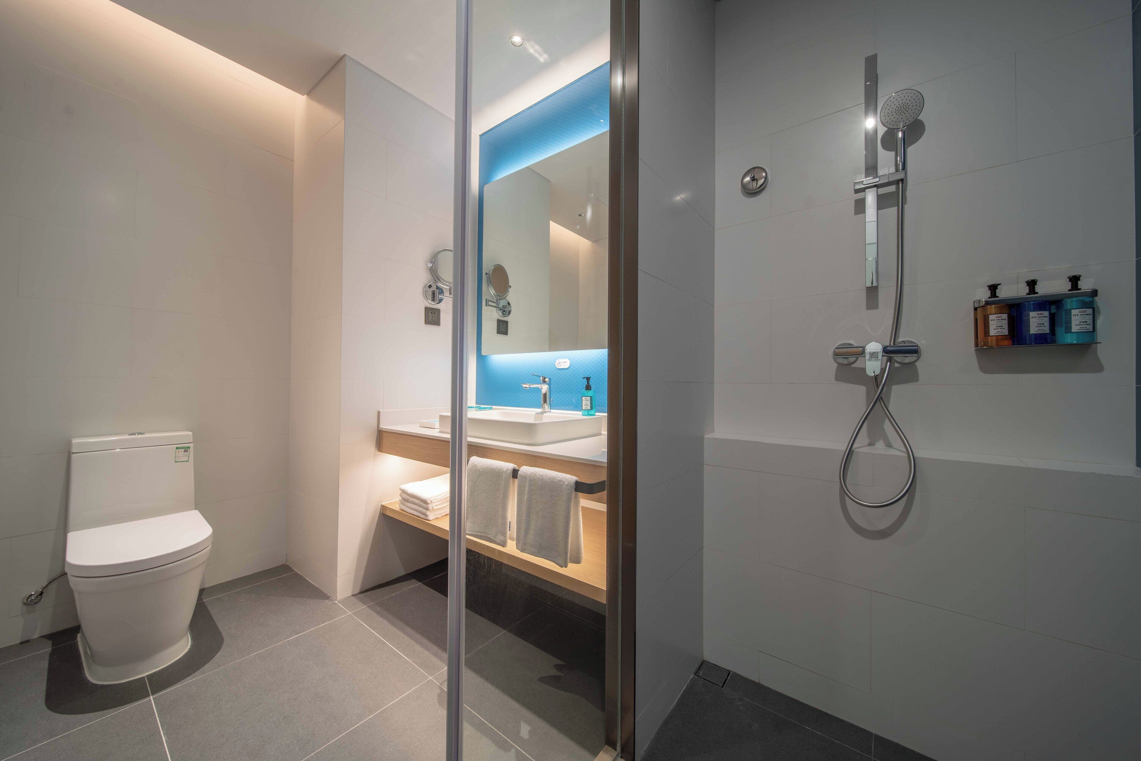 A modern bathroom with a toilet, a glass-enclosed shower area, a sink with a mirror, and a shelf with toiletries.