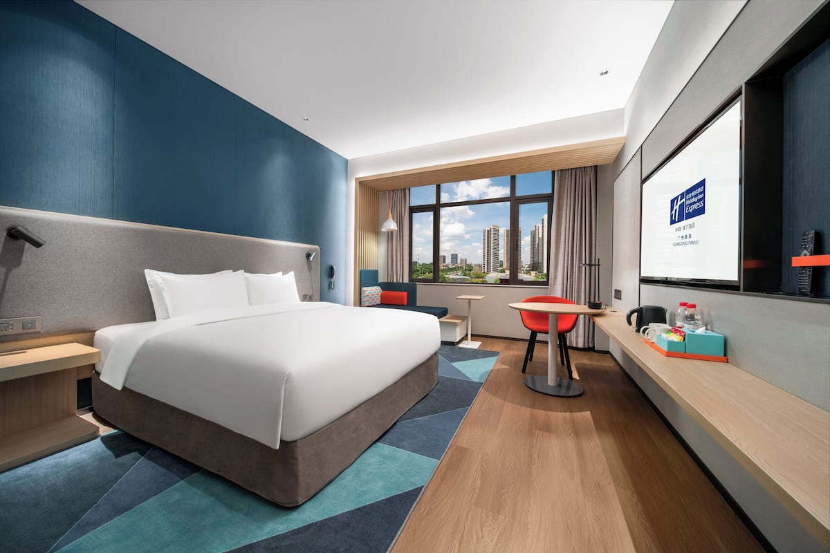 A modern hotel room with a large bed, a desk, a chair, and a view of the cityscape through large windows.