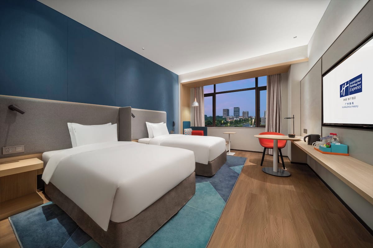 A modern hotel room with two beds, a desk, and a large window offering a city view.