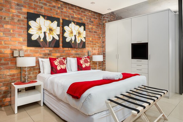 Habitación de hotel con pared de ladrillo, cama grande con ropa de cama blanca y detalles en rojo, televisión y armario.