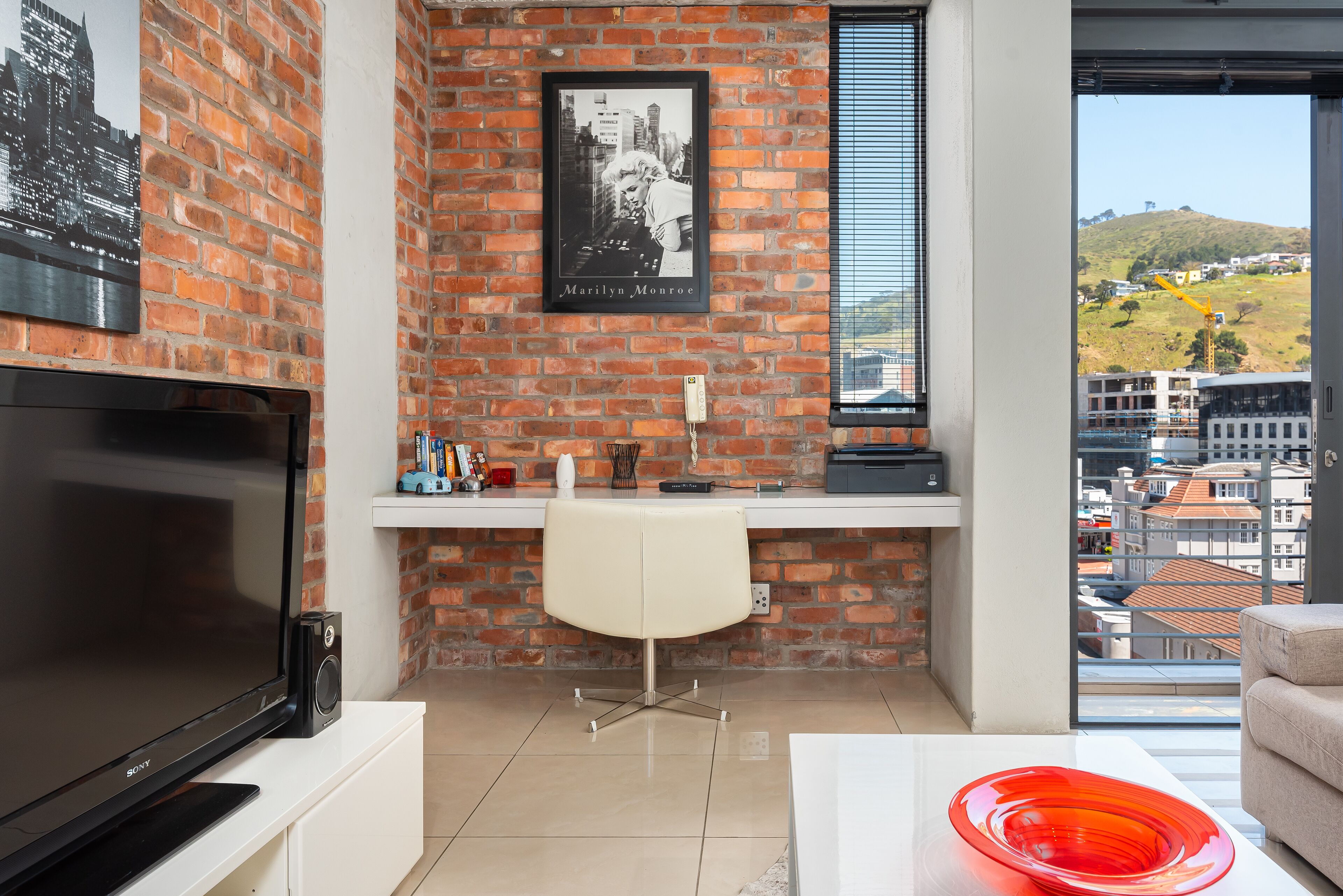 A modern living room with a brick wall, a flat-screen TV, a white desk, a white chair, and a red glass bowl on a white table.