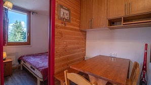 Quarto pequeno com uma mesa de madeira, uma cama e uma janela com vista para uma árvore.