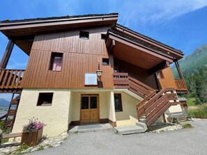 A two-story wooden chalet with a balcony, a wooden staircase, and a stone pathway leading to the entrance.