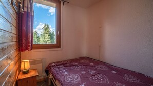 Um quarto pequeno e arrumado com uma cama de solteiro, uma janela com cortinas, um abajur e um radiador.