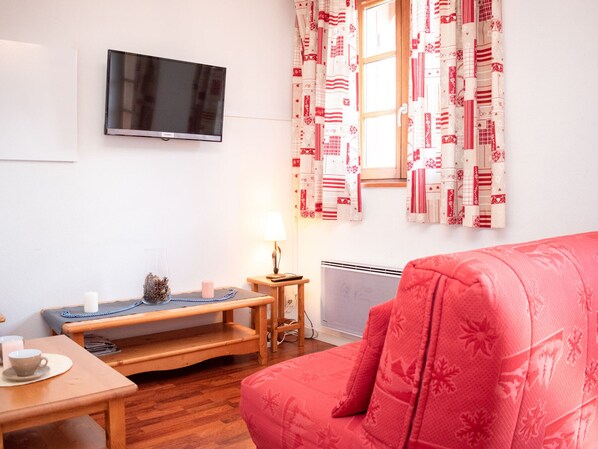 A cozy living room with a red sofa, a wooden bench, a TV mounted on the wall, and patterned curtains. - 2-Room Apartment with Balconies - Sleeps 6 (Vignec)