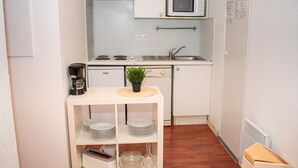 A compact kitchen with a microwave, stove, sink, and a small shelf with dishes and a plant.