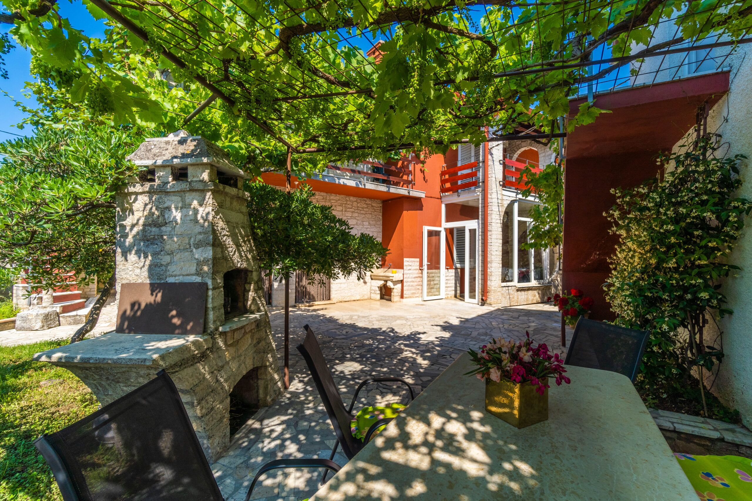 A stone structure with a bench and table, surrounded by greenery and a building with a red facade.