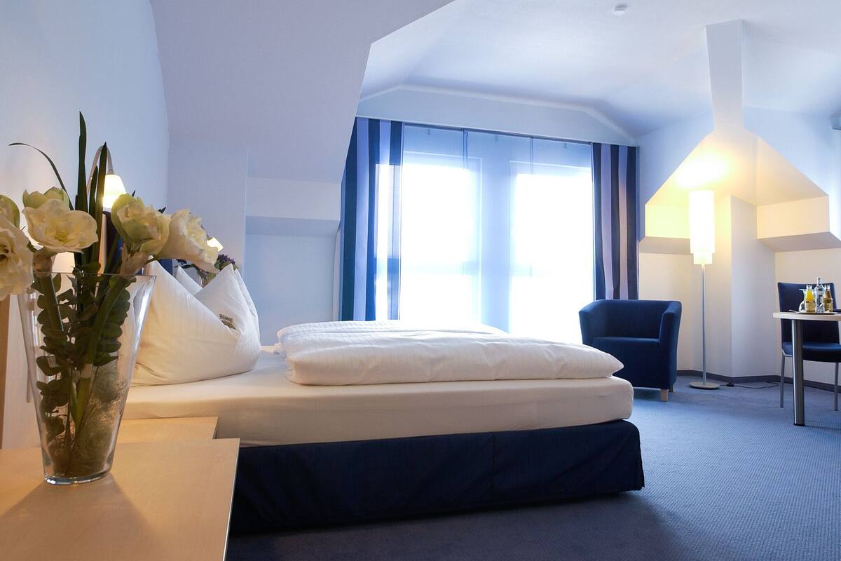 A hotel room with a bed, a chair, a desk, and a vase with flowers.