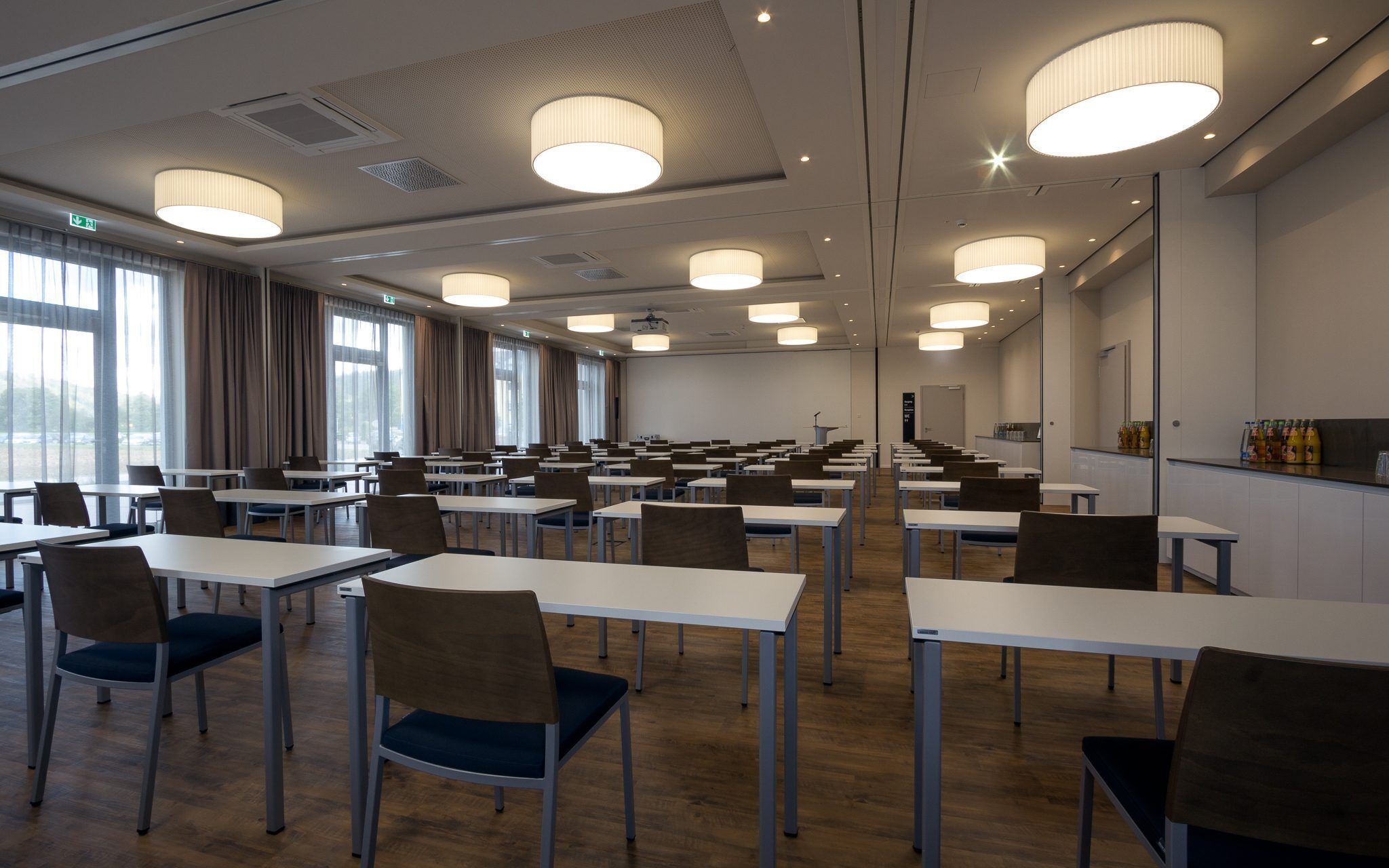 A conference room with tables and chairs, large windows, and ceiling lights.