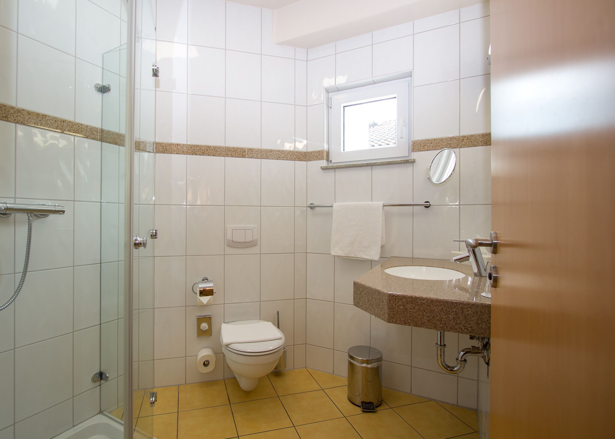 A bathroom with a glass-enclosed shower, a toilet, a sink with a granite countertop, and a window providing natural light.