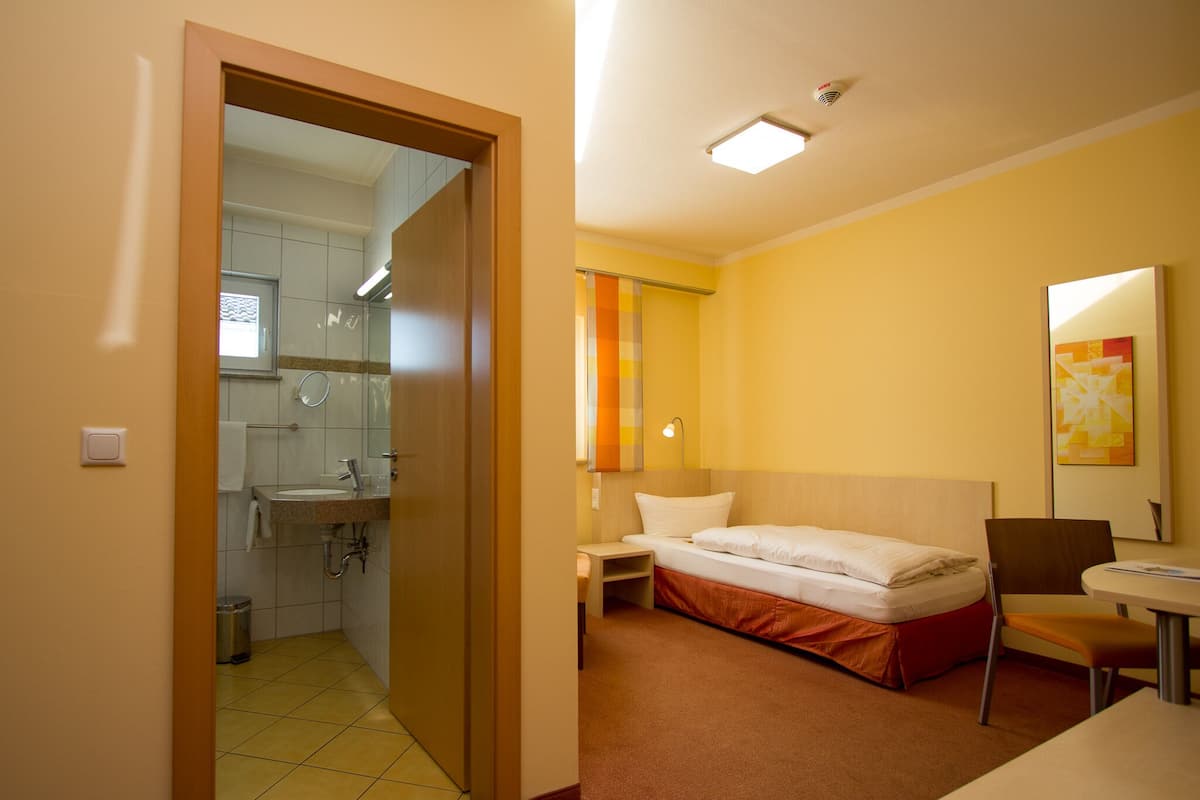 A hotel room with a bed, a desk, a chair, a small table, a mirror, and a bathroom with a sink and toilet.