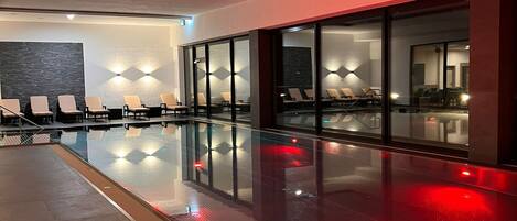 A modern indoor swimming pool with glass walls, lounge chairs, and ambient lighting.