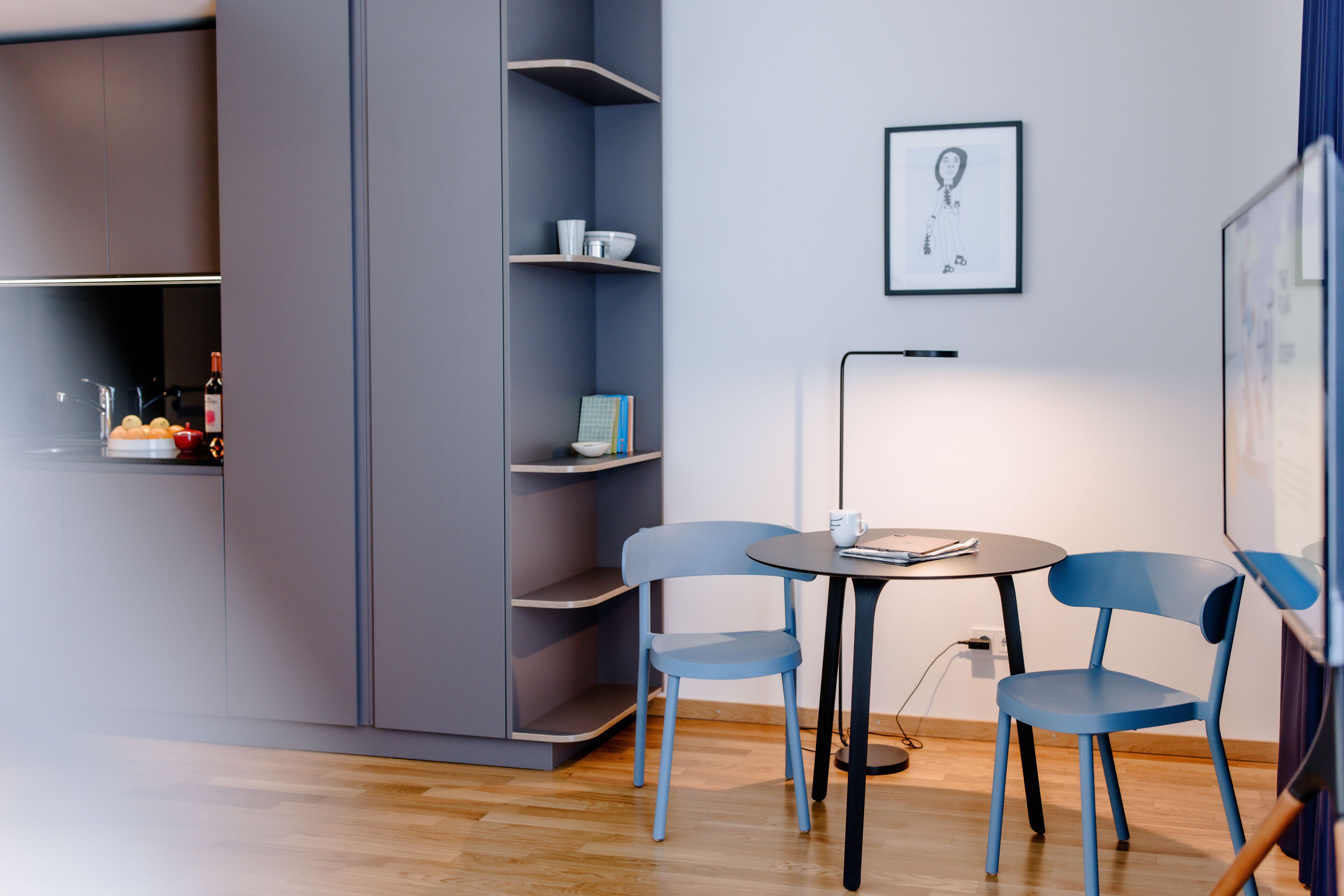 Une pièce moderne avec un meuble gris, une chaise bleue, une petite table ronde et un tableau encadré au mur.