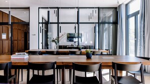 A modern dining area with wooden tables and black chairs, a potted plant, and a view through large windows. - THE FLAG Meiller Gärten (Munich)