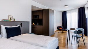 A modern hotel room with a large bed, a desk, and a kitchenette. - THE FLAG Meiller Gärten (Munich)