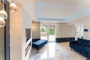 Een moderne woonkamer met een open haard, een blauwe bank en een groot raam met uitzicht op een tuin.
