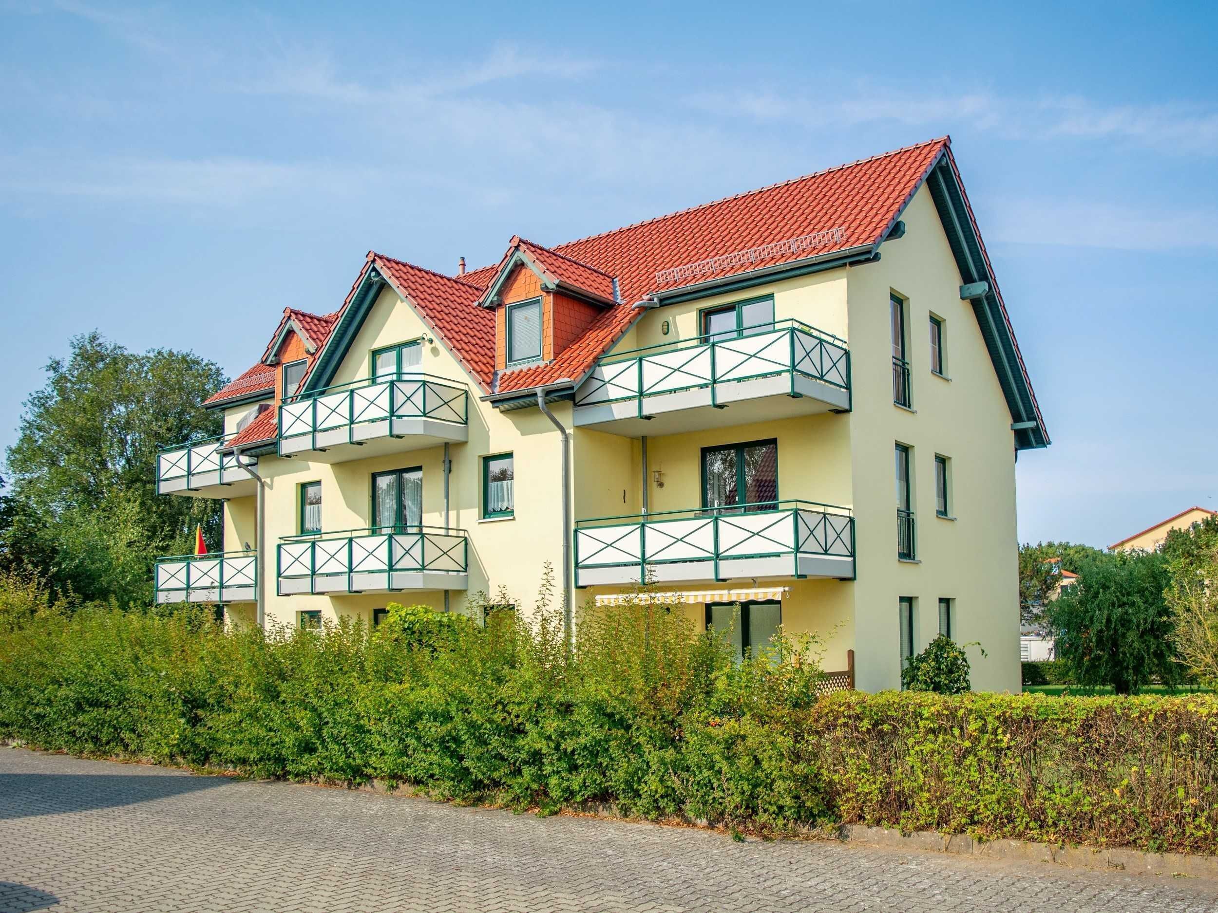 Ein mehrgeschossiges Wohngebäude mit Balkonen, einem roten Ziegeldach und einer gepflasterten Auffahrt.