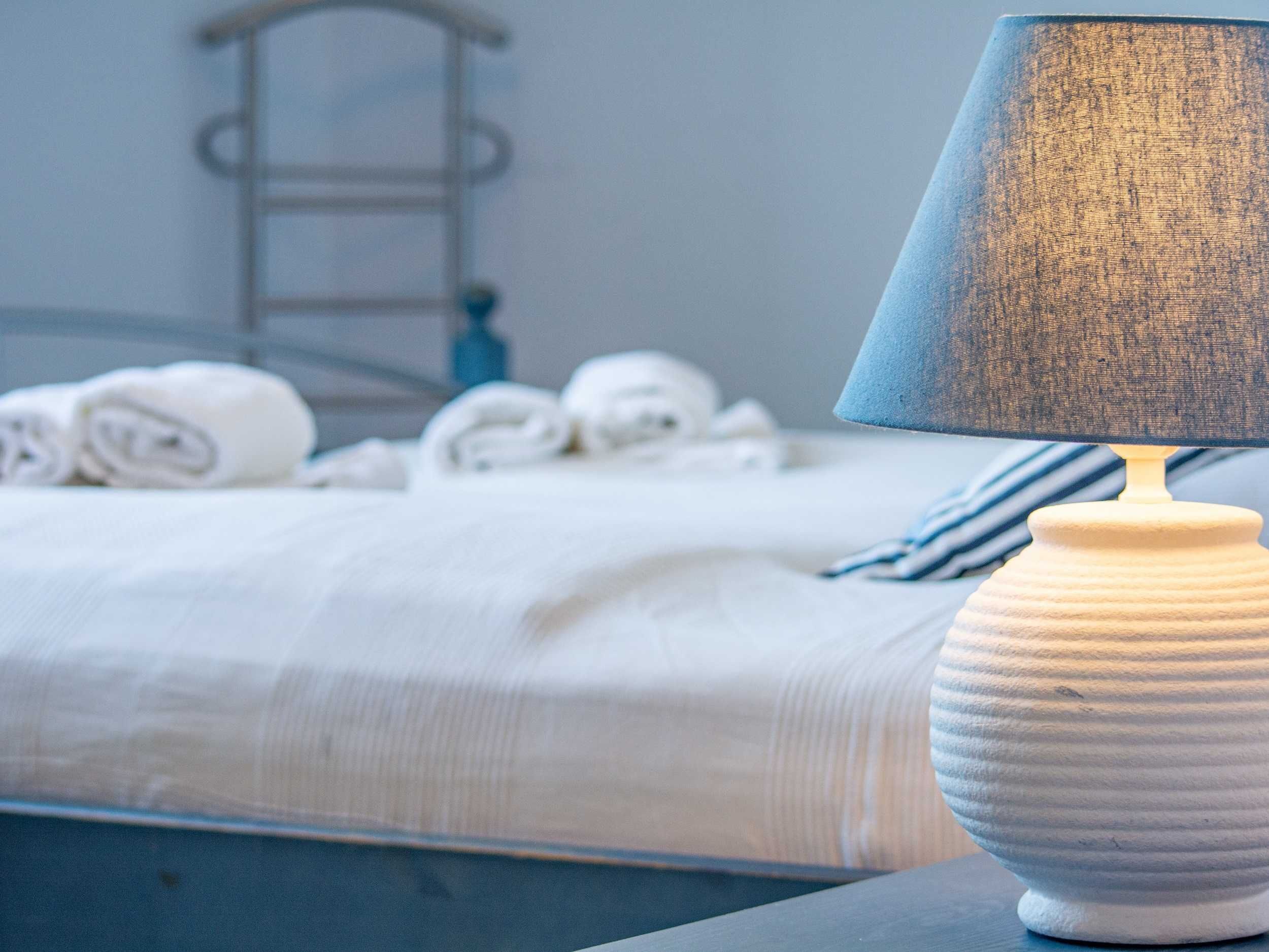 Een net opgemaakt bed met witte lakens en opgerolde handdoeken, een witte textuur lamp op een nachtkastje, en een metalen ladder die tegen de muur staat.