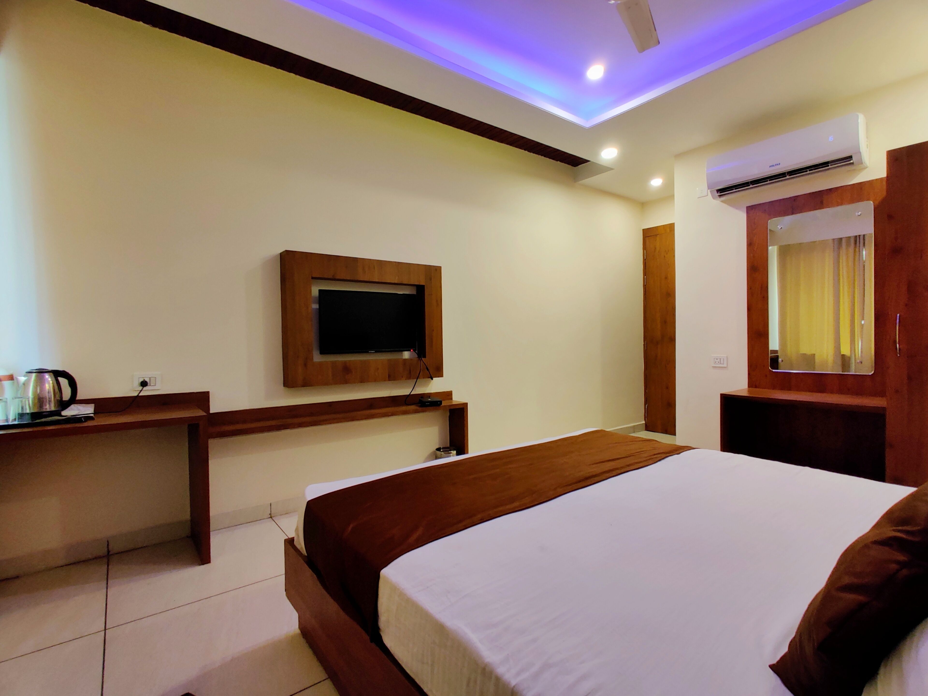 A hotel room with a bed, a TV mounted on the wall, an air conditioning unit, a wooden desk, and a mirror.