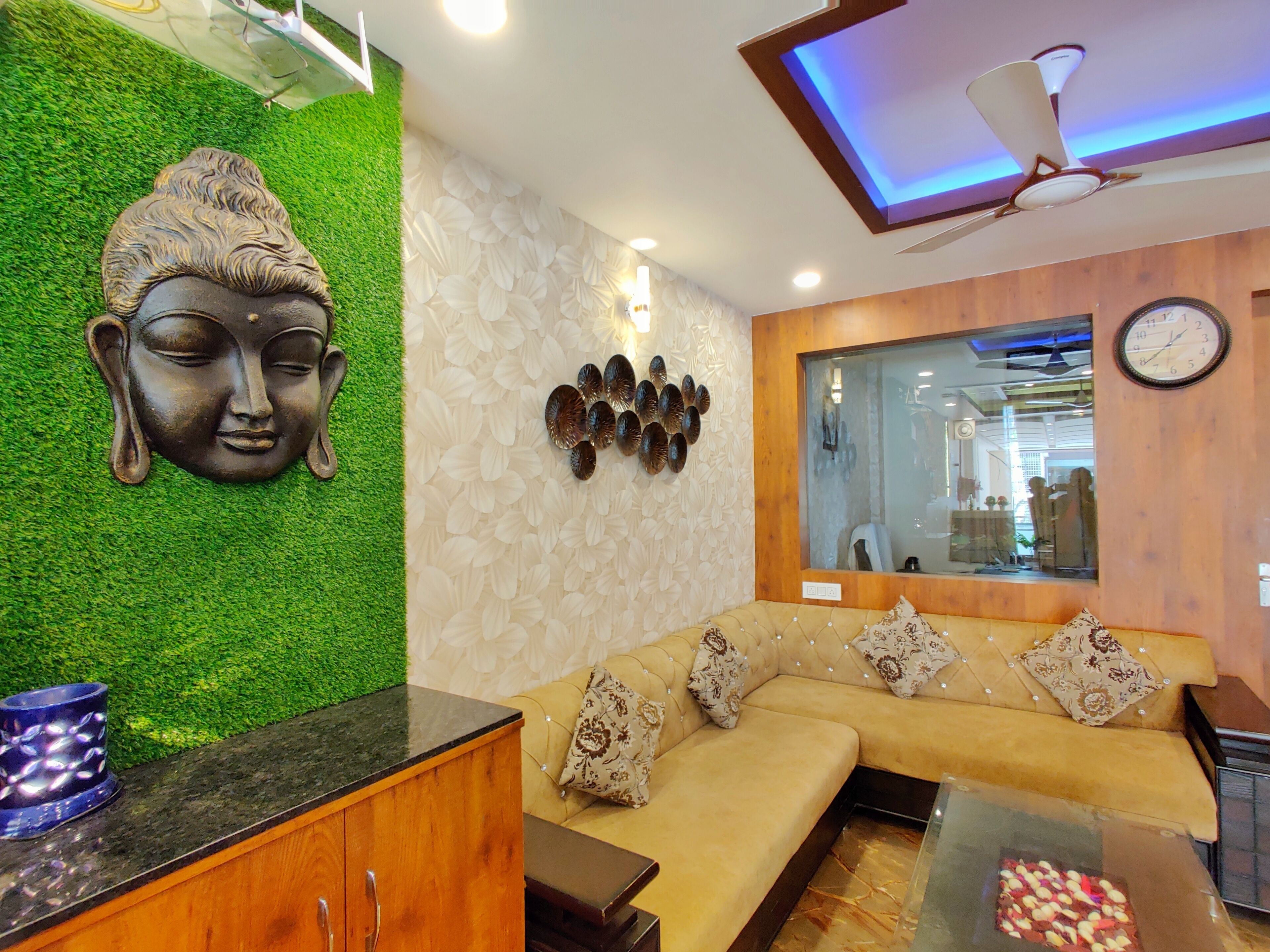 Ein modernes Wohnzimmer mit grüner Wand, einer Buddha-Kopf-Skulptur, einem hölzernen Deckenventilator, einer Couch mit gemusterten Kissen und einem Glastisch.