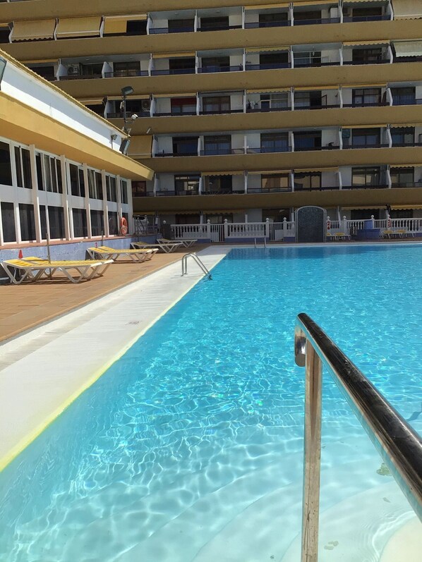 Una piscina con agua azul cristalina, rodeada por una terraza con sillas de descanso y un edificio con varios balcones al fondo.