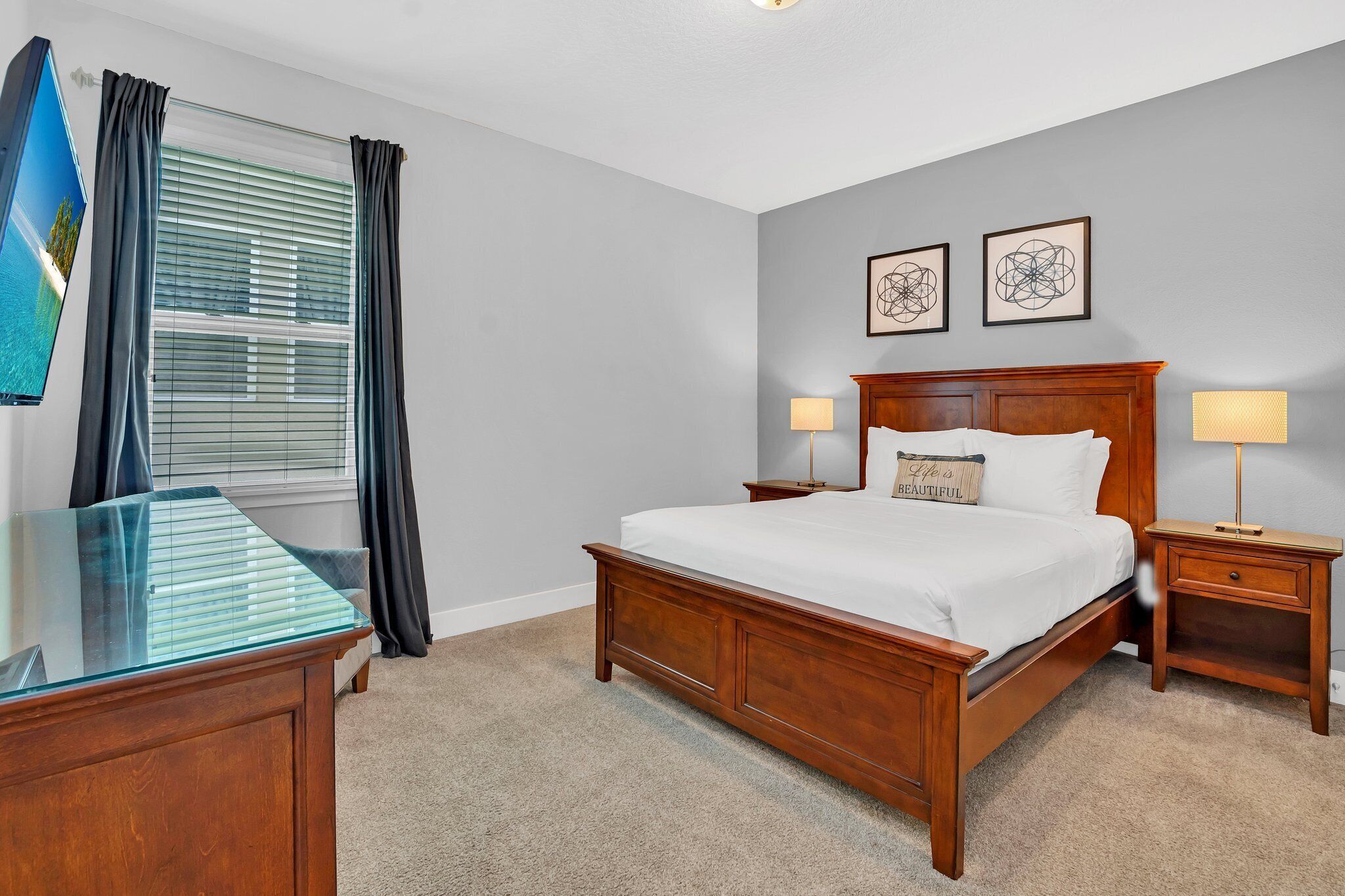 A bedroom with a wooden bed, two bedside tables with lamps, a large window with blinds, and a flat-screen TV on a stand.