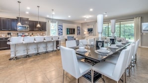A modern kitchen with dark cabinets, a central island, and a dining area with a long table and white chairs. - Villa W/ Pool, Movie Theater & Game Roomsleeps 20 (Kissimmee)