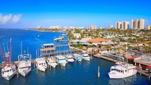 A marina with several boats docked, a pier, and a coastal cityscape with buildings and palm trees. - Oceanfront 1BR | Balcony | Steps to Sunglow Pier | Free Daily Activities Included (Daytona Beach Shores)