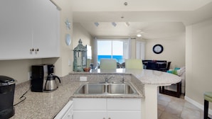 A kitchen with a granite countertop, stainless steel sink, and white cabinets. There is a coffee maker and a kettle on the counter.