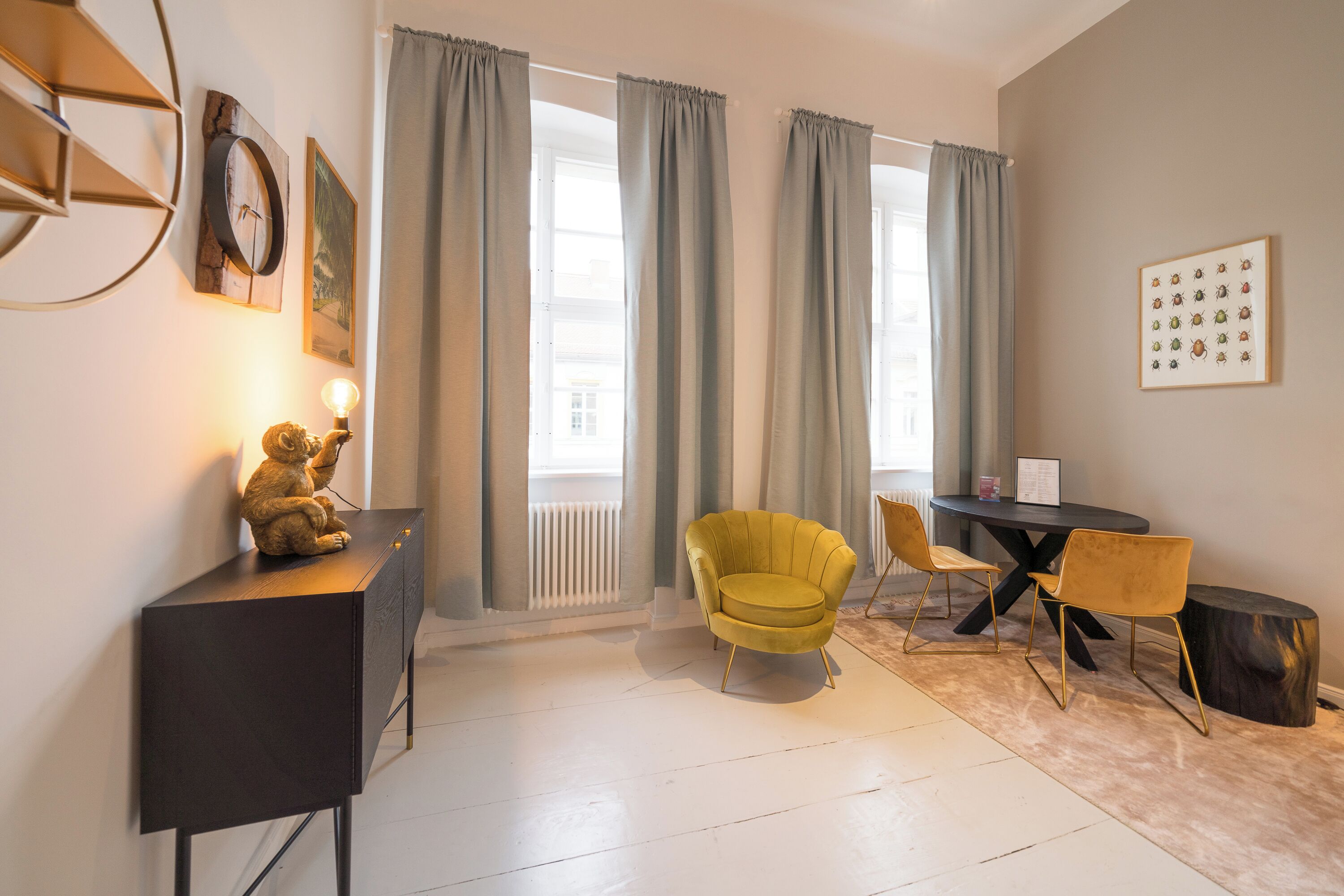 A modern living room with a sofa, a small table, and a chair. There is a window with curtains, a radiator, and a wall clock.