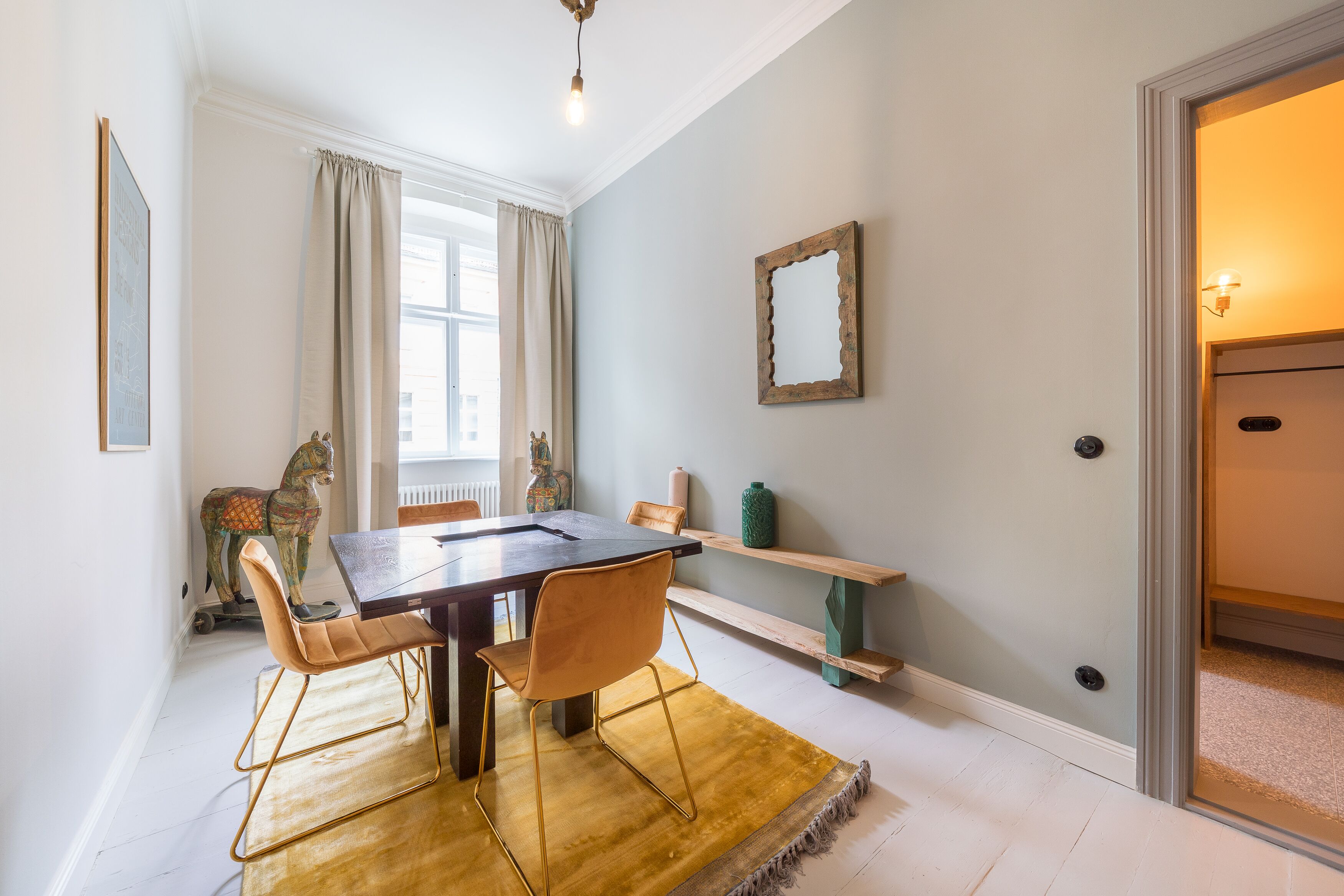 A dining area with a wooden table, two chairs, a yellow rug, a horse sculpture, and a mirror on the wall.