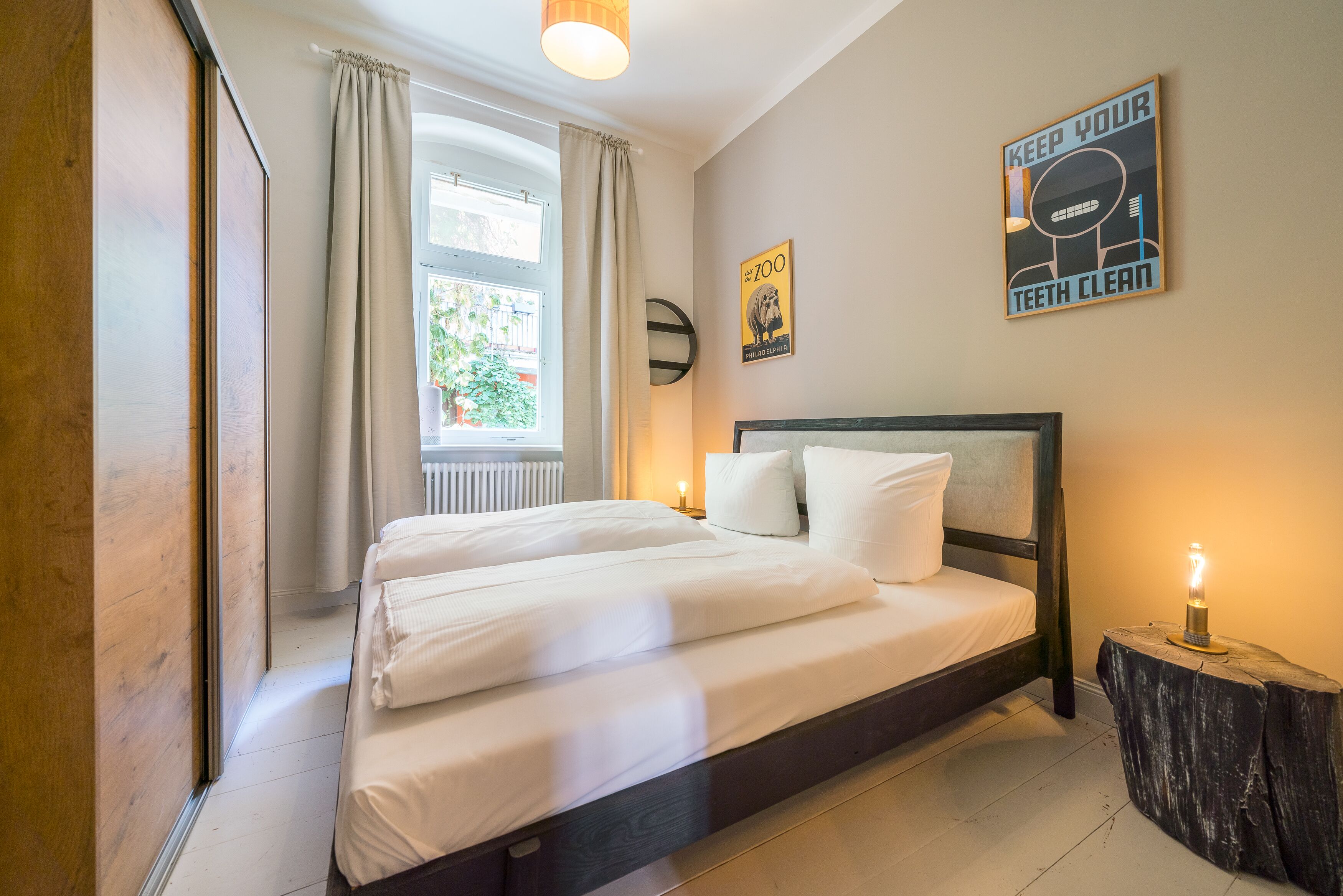 A bedroom with a bed, a bedside table with a lamp, a window with curtains, and a poster on the wall.