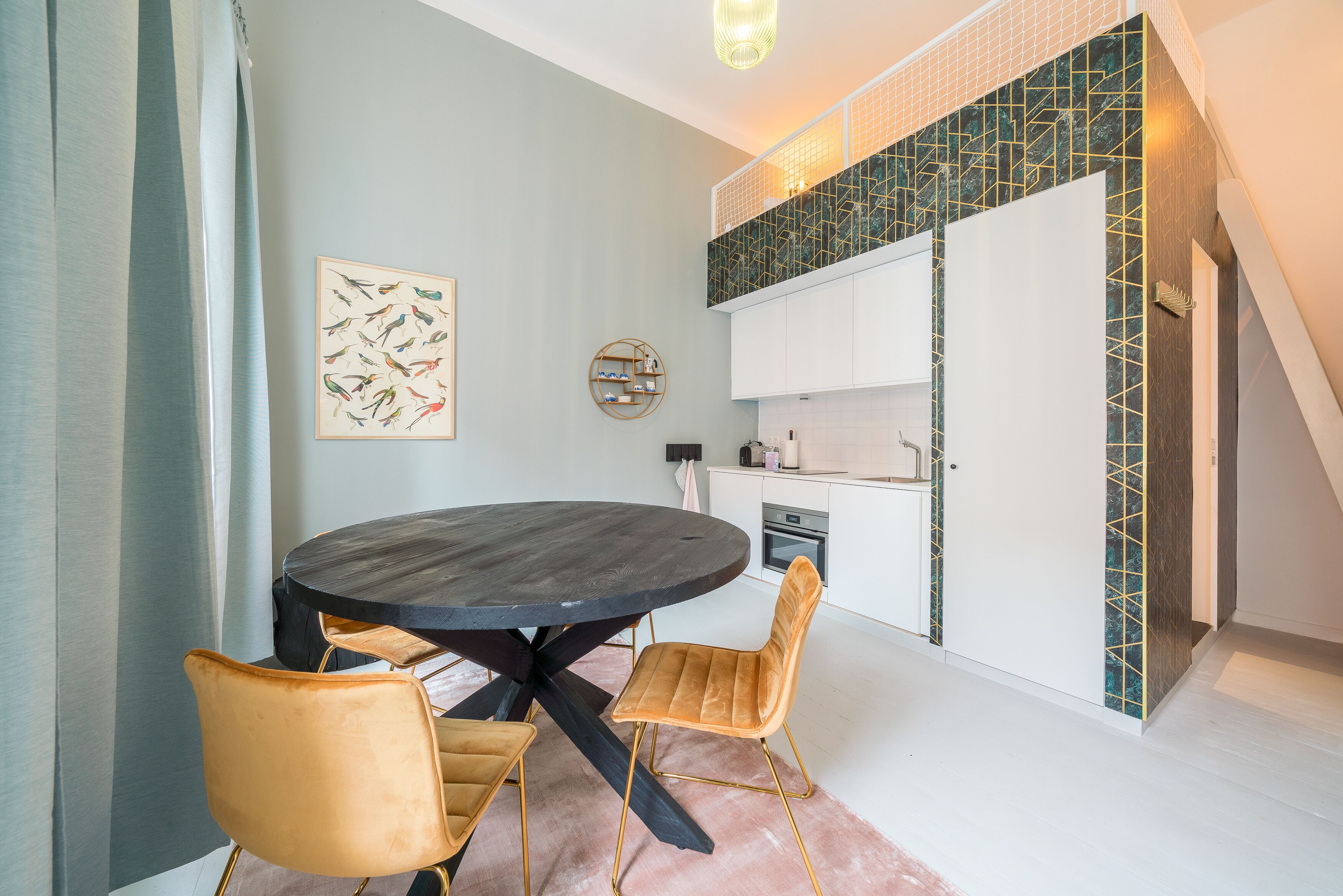 Un coin repas avec une table ronde et deux chaises, une cuisine avec des meubles blancs et un mur décoratif orné d’un motif géométrique.