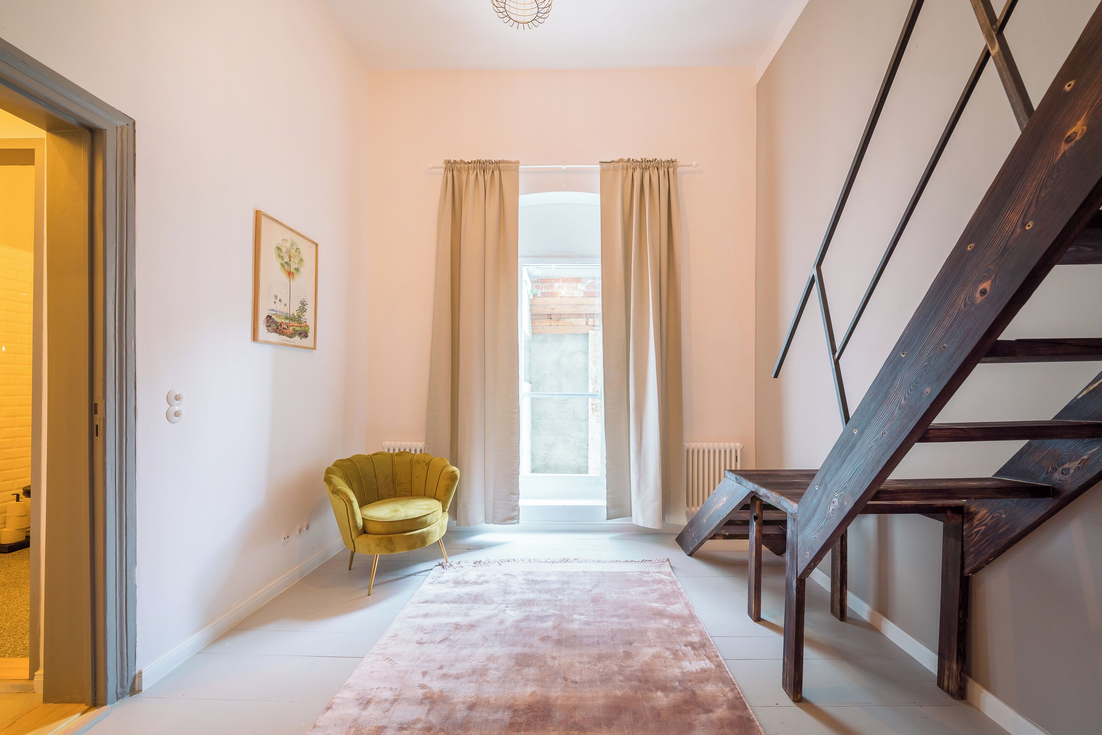 A hallway with a staircase, a yellow chair, a pink rug, and a window with curtains.