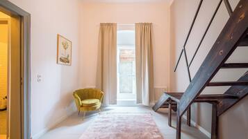 A hallway with a staircase, a yellow chair, a pink rug, and a window with curtains.