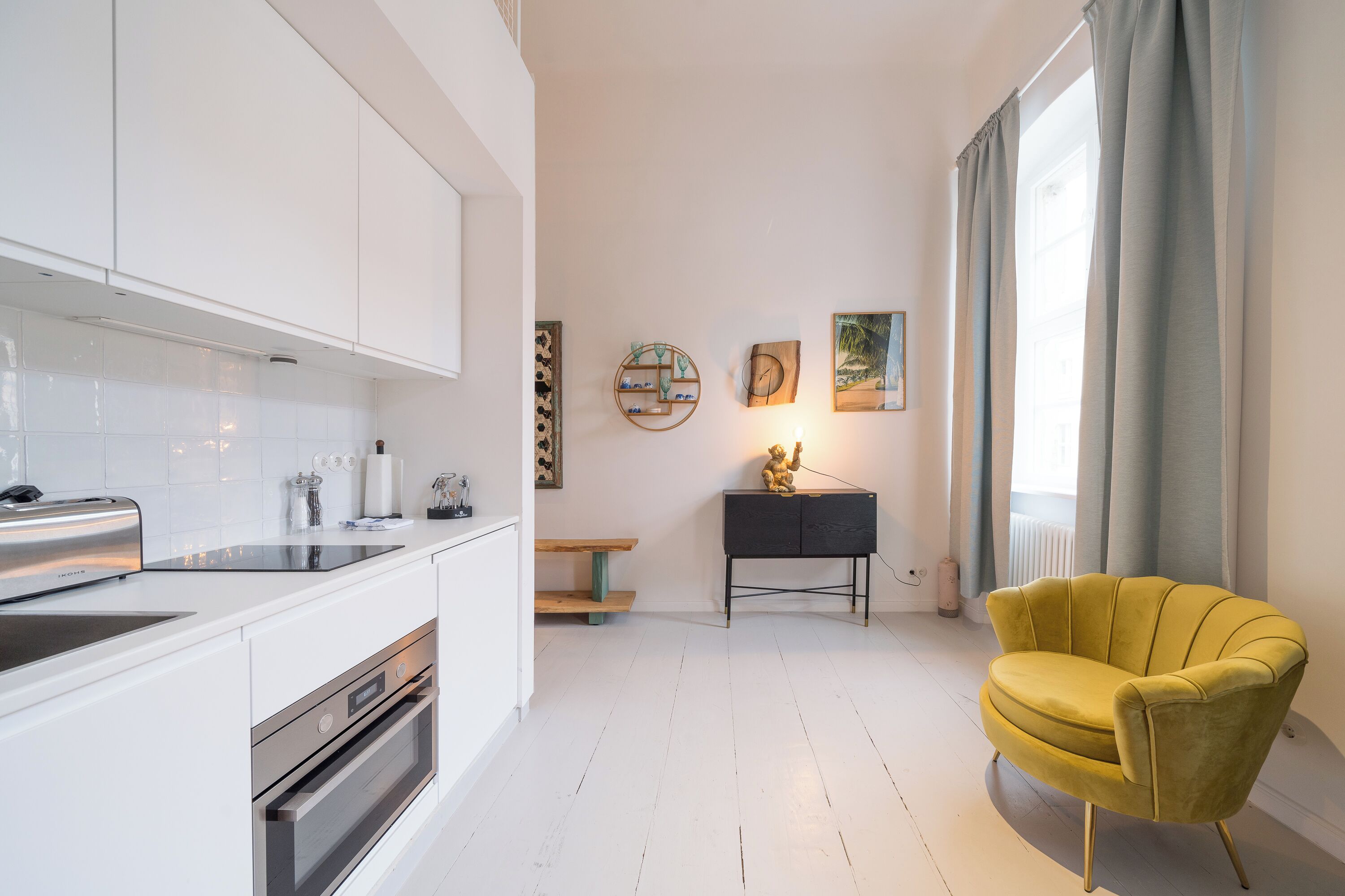 Eine moderne Küche mit weißen Schränken, einem Einbauofen und einem Toaster. Es gibt eine kleine Holzbank und einen schwarzen Beistelltisch mit einem dekorativen Gegenstand. Ein gelber Sessel steht in der Nähe eines großen Fensters mit Vorhängen.