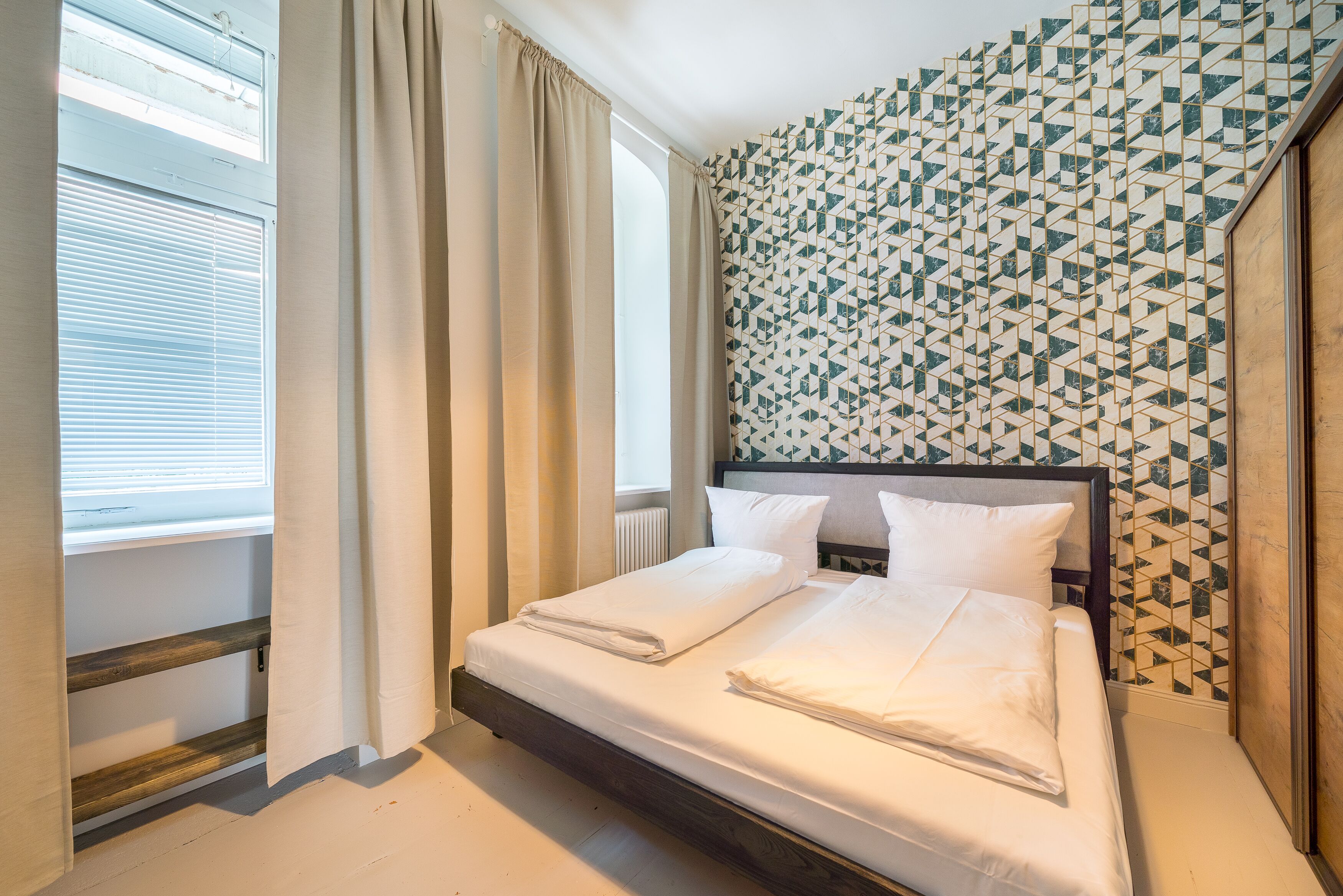 Una habitación de hotel con cama, cortinas, ventana y un papel tapiz estampado.