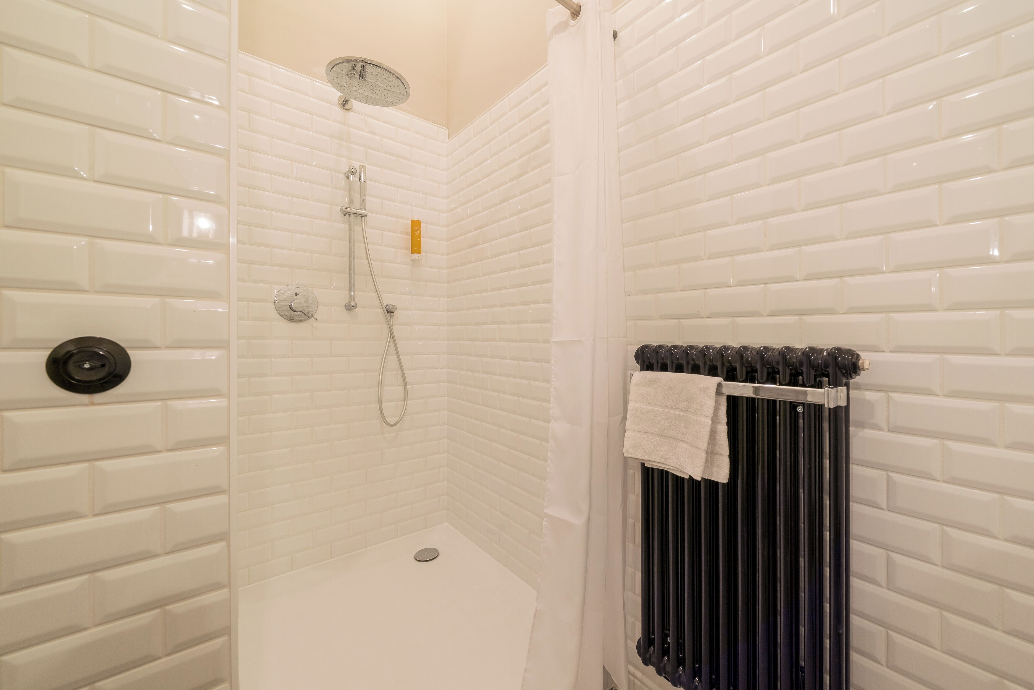 Een douche met witte betegelde wanden, een zwarte radiator en een witte handdoek aan de radiator.