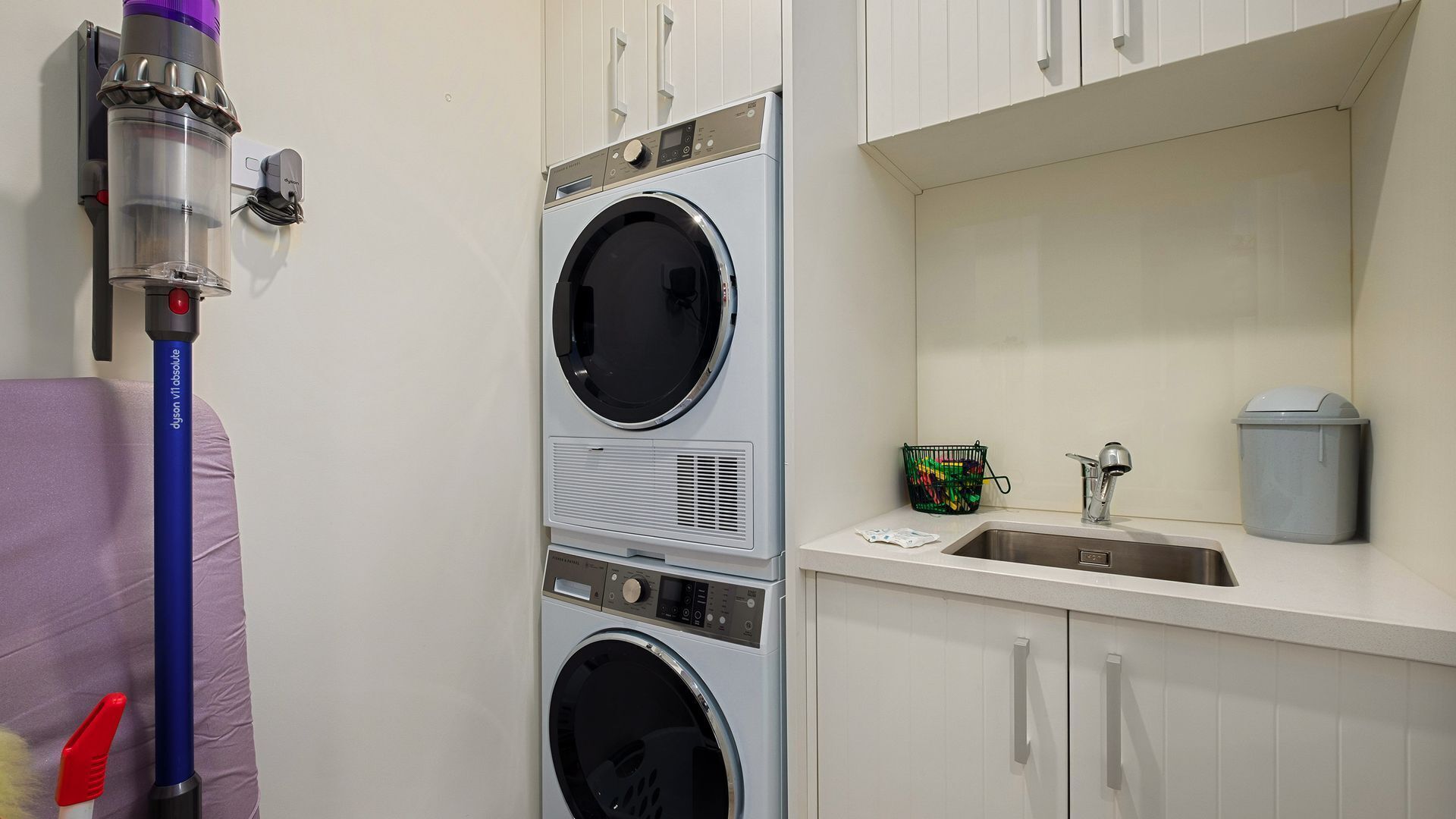 A laundry room with two stacked front-loading washing machines, a Dyson vacuum cleaner, a sink with a trash can, and a basket of laundry.